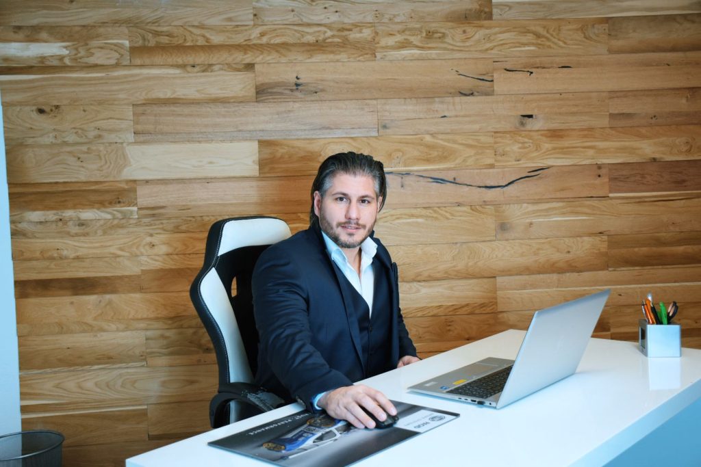 Das Bild zeigt Jürgen Pichler, in einem dunkelblauen Anzug und weisen Hemd. Jürgen sitzt auf einem schwarz weißen Bürostuhl vor einem weißen Schreibtisch. Beide Hände liegen am weisen Schreibtisch auf, wo ein grauer Laptop, eine schwarze Maus und ein Mauspad zusehen sind. Im Hintergrund ist eine Holzwand aus Eiche zusehen.
